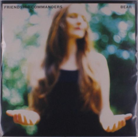 Text oben: "FRIENDSHIP COMMANDERS" links, "BEAR" rechts. Unscharfe Frau mit langen Haaren, die Hände geöffnet.