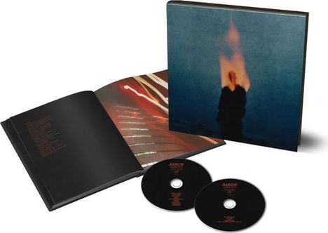 „Amor“ auf zwei CDs; links ein aufgeschlagenes Buch mit Texten; im Hintergrund eine mysteriöse Abbildung.