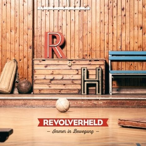 „REVOLVERHELD - Immer in Bewegung“ auf rotem Banner. Holzkiste mit Buchstaben „R“ und „H“, Turnhallenatmosphäre.