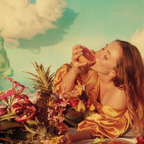 Ich sehe keinen Text. Eine Frau in gelbem Kleid genießt eine halbe Grapefruit, umgeben von exotischen Früchten und Blumen.