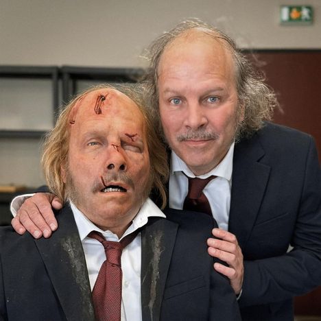 Zwei Männer in Anzügen. Einer hat Verletzungen im Gesicht. Sie stehen nah beieinander in einem Raum.