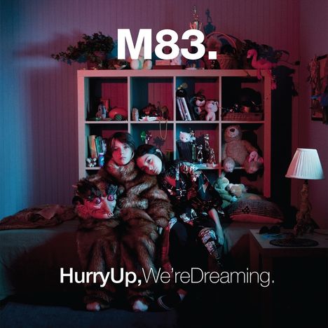 "M83." und "Hurry Up, We're Dreaming." Zwei Personen in einem fantasievollen, plüschigen Raum mit Spielzeug.