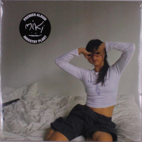 Logo mit Text "PREMIER ALBUM MIKI INDUSTRY PLANT". Eine Frau in weißem Oberteil und schwarzen Shorts sitzt auf einem Bett.