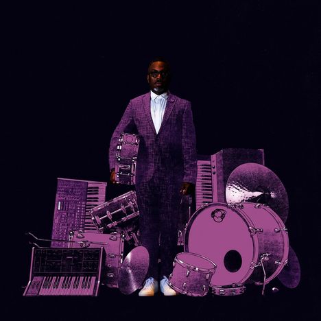 Kein lesbarer Text. Eine Person in Anzug, umgeben von lila Musikinstrumenten, darunter Keyboards und Schlagzeug.