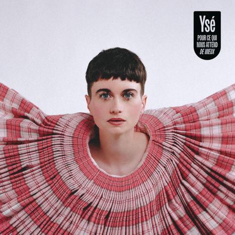 Text: „YsÉ POUR CE QUI NOUS ATTEND DE MIEUX“. Frau mit kurzem Haar trägt ein auffälliges, plissiertes Kleid in Rosa- und Rottönen.