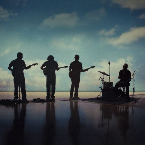 Ich kann keine Texte erkennen. Silhouette von vier Musikern mit Instrumenten vor einem Hintergrund aus blauen Wolken.