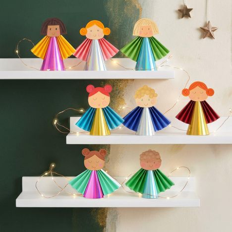 Sechs bunte Engelsfiguren aus Papier stehen auf weißen Regalbrettern, dekoriert mit Lichterketten und Sternen an der Wand.