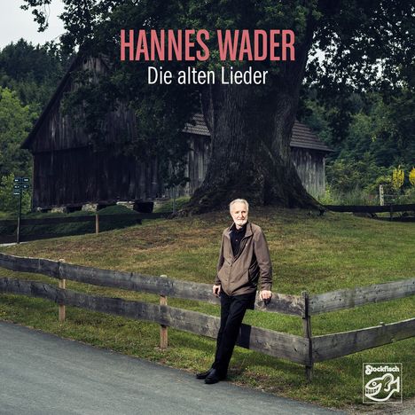 „HANNES WADER Die alten Lieder“. Ein älterer Mann lehnt an einem Holzzaun vor einem großen Baum und einer Scheune.