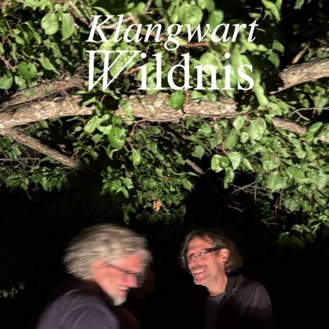 Text: "Klangwart Wildnis." Zwei Männer stehen unter einem belaubten Baum, einer lächelt, der andere ist unscharf.