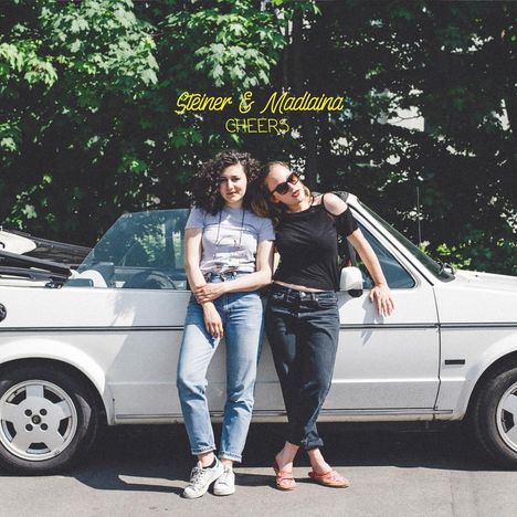 „Steiner & Madlaina CHEERS“ in gelber Schrift. Zwei Frauen lehnen lässig an einem weißen Cabrio vor grünen Bäumen.