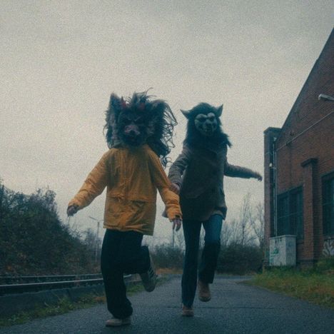 Zwei Personen laufen Hand in Hand, tragen Tiermasken und Jacken, links gelb, rechts braun, in industrieller Umgebung.