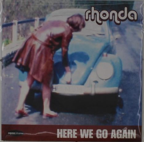 Texts: "rhonda" oben rechts, "HERE WE GO AGAIN" unten. Eine Person in einem braunen Outfit beugt sich zu einem blauen Auto.