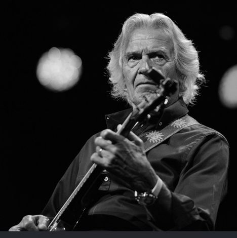 Ein älterer Mann spielt Gitarre, sein Gesichtsausdruck ist konzentriert. Er trägt ein Hemd mit Verzierungen am Kragen und Ärmel.