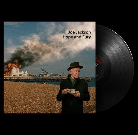 "Joe Jackson Hope and Fury." Eine Person im Mantel und Hut trinkt Tee, im Hintergrund brennt ein Pier.