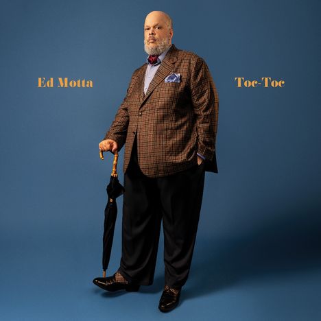 "Ed Motta" links, "Toe-Toc" rechts. Ein Mann im Anzug mit Regenschirm steht vor blauem Hintergrund.
