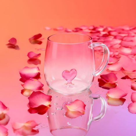 Ein durchsichtiger Becher mit Herzform im Boden, umgeben von rosa Blütenblättern auf einer rosa reflektierenden Oberfläche.