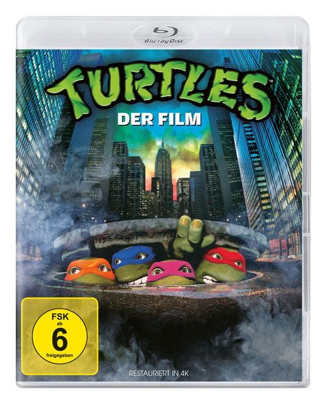 "TURTLES DER FILM", vier maskierte Schildkröten schauen aus einem Kanaldeckel hervor; Hochhäuser im Hintergrund.