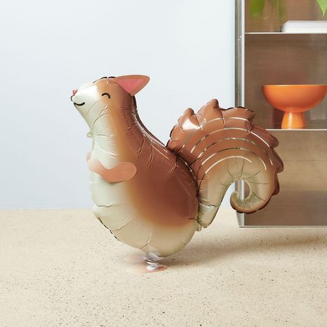 Eine Eichhörnchen-Heliumballonfigur steht aufrecht auf hellem Boden. Eine metallene Regalwand mit orangefarbener Schale ist daneben.