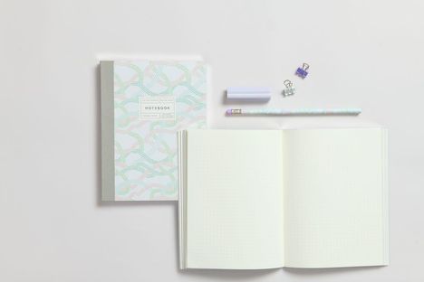 Ein Notizbuch mit Punktmuster, Bleistift, zwei Klammern und einer zylindrischen Box auf hellgrauem Hintergrund.