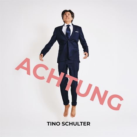 Text: "ACHTUNG" in großem Rot darüber "TINO SCHULTER". Ein Mann im Anzug schwebt mit nackten Füßen vor weißem Hintergrund.