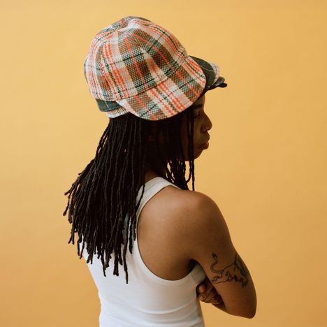 Person trägt einen karierten Hut, hat lange Dreadlocks, ein weißes Oberteil und ein Tattoo auf dem Arm. Hintergrund ist gelb.