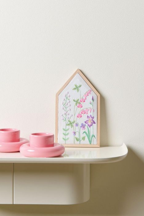 Keine Texte erkennbar. Eine Illustration von Blumen in einem holzgerahmten Hausform mit zwei rosa Kerzenhaltern daneben.