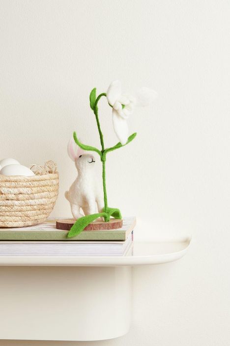 Ein Filzhase neben einem Korb mit Eiern, hält eine weiße Blume. Alles auf gestapelten Büchern vor weißem Hintergrund.