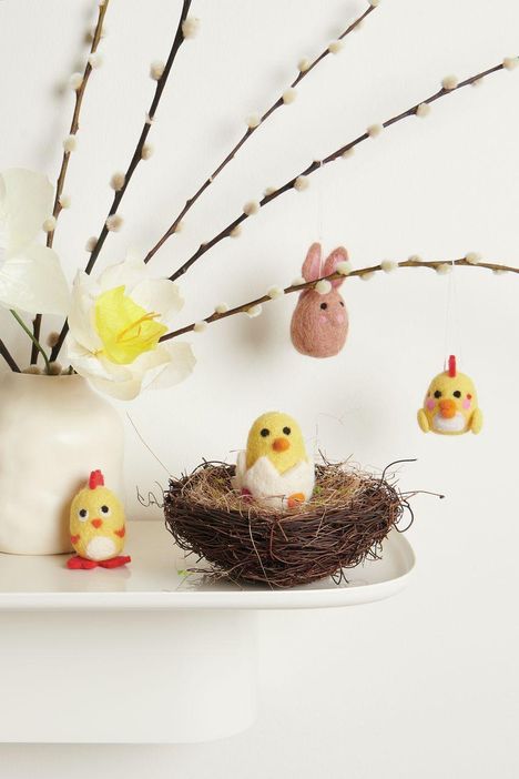 Kükenfiguren aus Filz, sitzend und hängend, umgeben von Zweigen und Blumen in einer weißen Vase auf einem Regal.