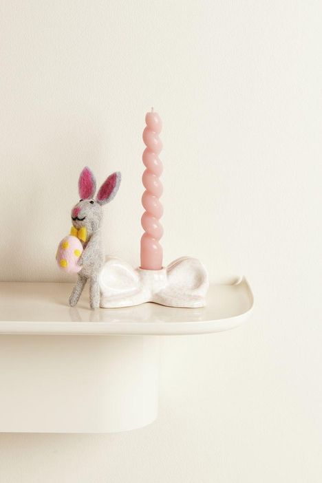 Ein grauer Hase mit gelber Fliege hält ein gepunktetes Ei. Daneben eine rosa Wendekerze in einem Kerzenständer.