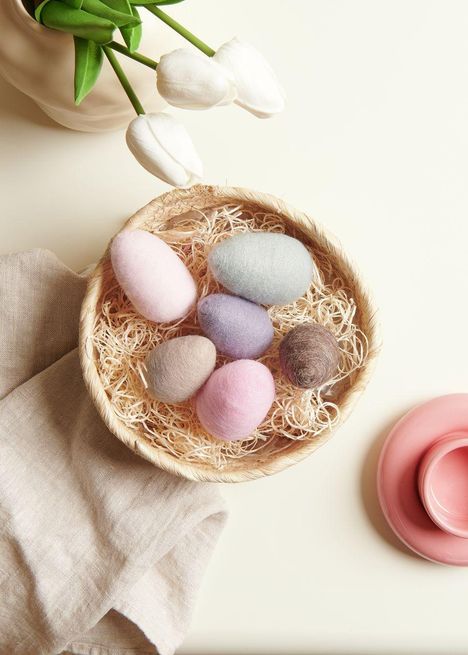 Sechs pastellfarbene Filzeier in einem Korb mit Stroh; daneben weiße Tulpen und eine rosa Tasse.