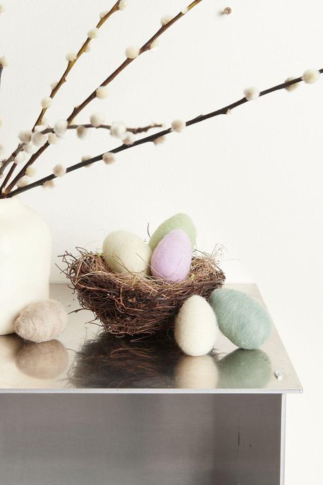 Einige pastellfarbene Filzeier liegen in einem Nest auf einem Tisch neben einer weißen Vase mit Weidenkätzchen.