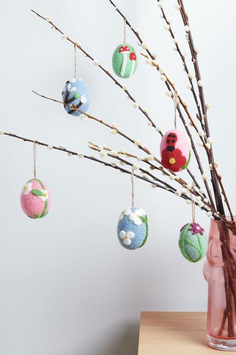 Sechs bunte Filzeier mit Blumen- und Pilzmuster hängen an einem Weidenzweig in einer rosa Vase.