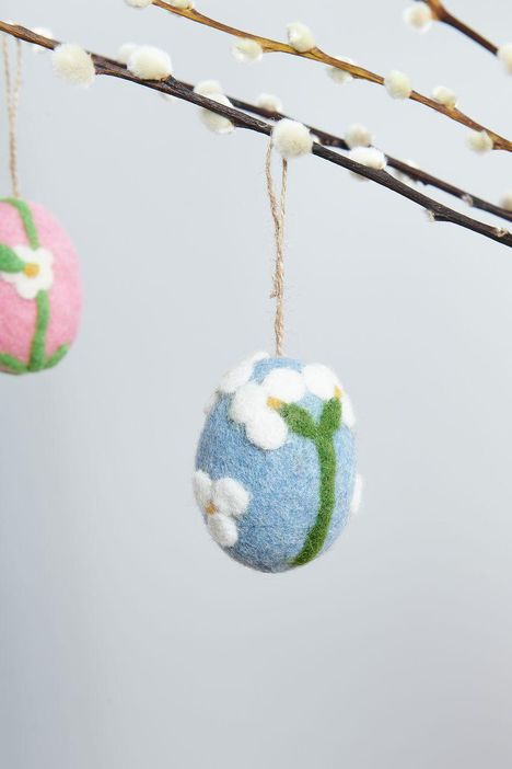 Blaue Filzkugel mit weißen Blüten hängt an einem Ast mit flauschigen Knospen. Links eine rosa Kugel sichtbar.