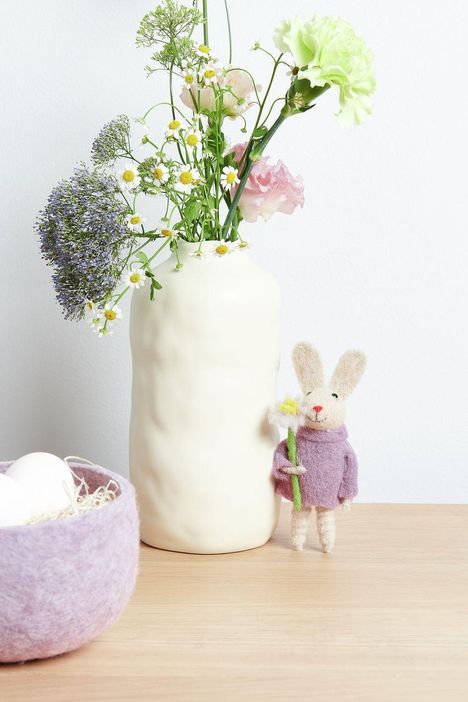 Kein Text zu sehen. Eine Vase mit bunten Blumen und daneben steht ein Stoffhase mit lila Pullover, der eine Blume hält.