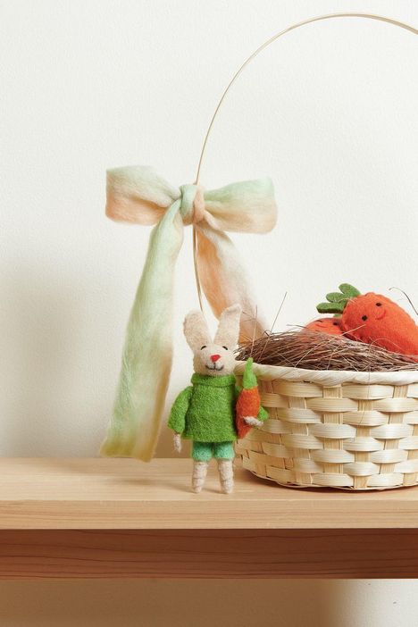 Ein kleiner Filzhase in grünem Outfit hält eine Karotte, daneben ein Korb mit Filzkarotten und einer bunten Schleife.