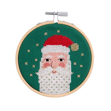 Ein Stickrahmen zeigt eine Kreuzstich-Illustration von Santa Claus mit roter Mütze auf grünem Hintergrund und goldenen Sternen.