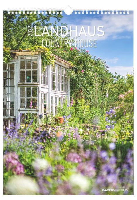 2024 LANDHAUS COUNTRY HOUSE. Ein weißes Glashaus im üppigen Garten mit bunten Blumen, umgeben von Bäumen und Sträuchern.