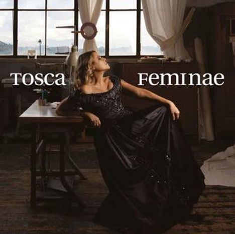 Text "tosca feminae". Eine Frau in schwarzem Kleid lehnt an einem Tisch in einem industriellen Raum mit großen Fenstern.