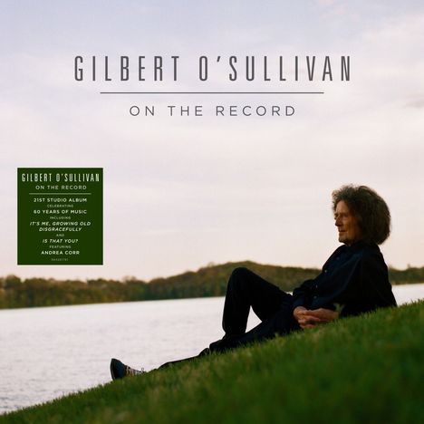 "GILBERT O'SULLIVAN ON THE RECORD. Person sitzt auf einem Hügel am Wasser, im Hintergrund Bäume."