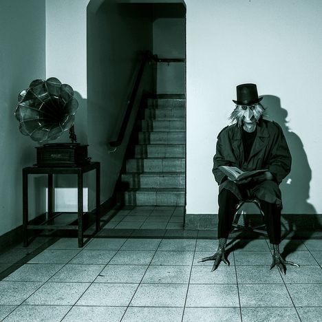 Ein Raum mit Schallplattenspieler, Treppe im Hintergrund. Person mit Vogelmaske und Zylinder sitzt lesend auf einem Stuhl.