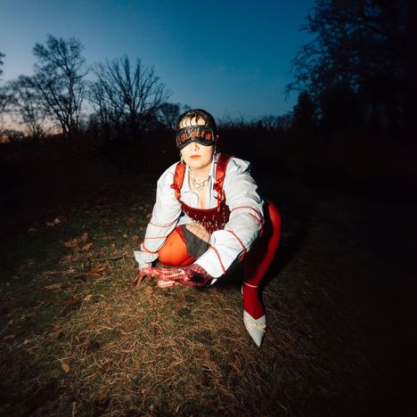 Person in modischem Outfit und Maske mit Schriftzug „Empower“ kauert auf Gras; Hintergrund: Dämmerung, Bäume.