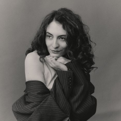 Ein schwarz-weiß Porträt zeigt eine Frau mit lockigem Haar, die nachdenklich lächelt und ihre Hände unter ihr Kinn legt.