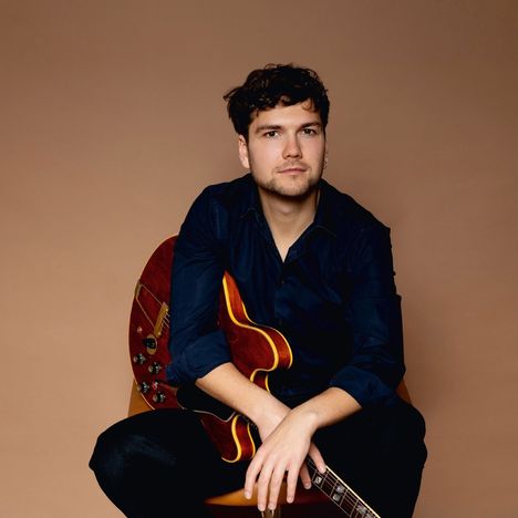 Ein Mann in dunklem Hemd sitzt mit einer braunen Gitarre vor einem neutralen, braunen Hintergrund.