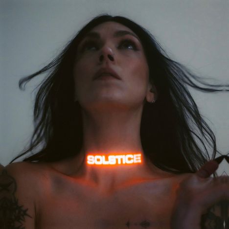 Der Text „SOLSTICE“ leuchtet am Hals einer Person mit langen dunklen Haaren und einigen Tattoos auf den Schultern.