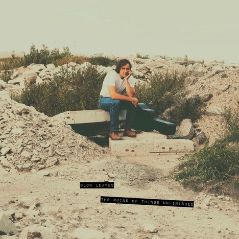  Texte: "SLOW LEAVES", "THE RUINS OF THINGS UNFINISHED". Person sitzt nachdenklich auf Betonblöcken in Trümmern.