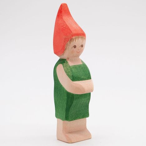 Eine einfache, hölzerne Figur im grünen Outfit mit roter, spitzer Mütze, lächelt freundlich, Arme verschränkt.