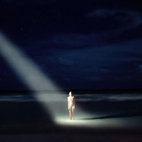 Ein heller Lichtstrahl fällt auf eine stehende Person nachts am Strand, der Himmel ist sternenklar und dunkel.