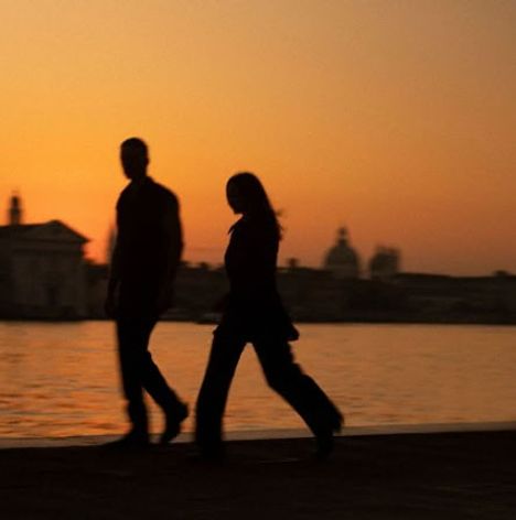 Keine sichtbaren Texte. Zwei Personen gehen am Wasser entlang, Silhouetten vor einem orangefarbenen Sonnenuntergang.