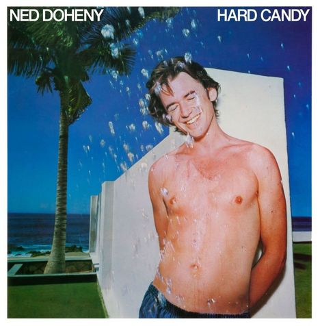 "NED DOHENY" und "HARD CANDY" oben. Ein lächelnder, oberkörperfreier Mann mit Wasserstrahlen, Palme und Meer im Hintergrund.