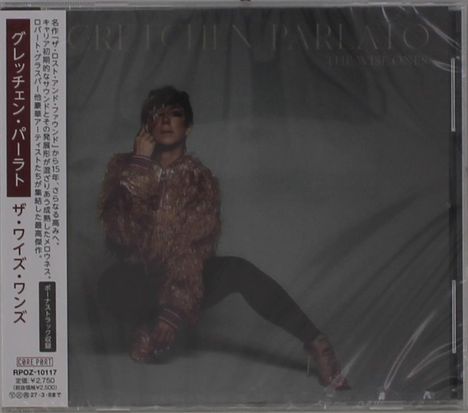 Auf der linken Seite vertikal japanischer Text. In der Mitte eine Person in einem flauschigen Outfit vor dunklem Hintergrund.